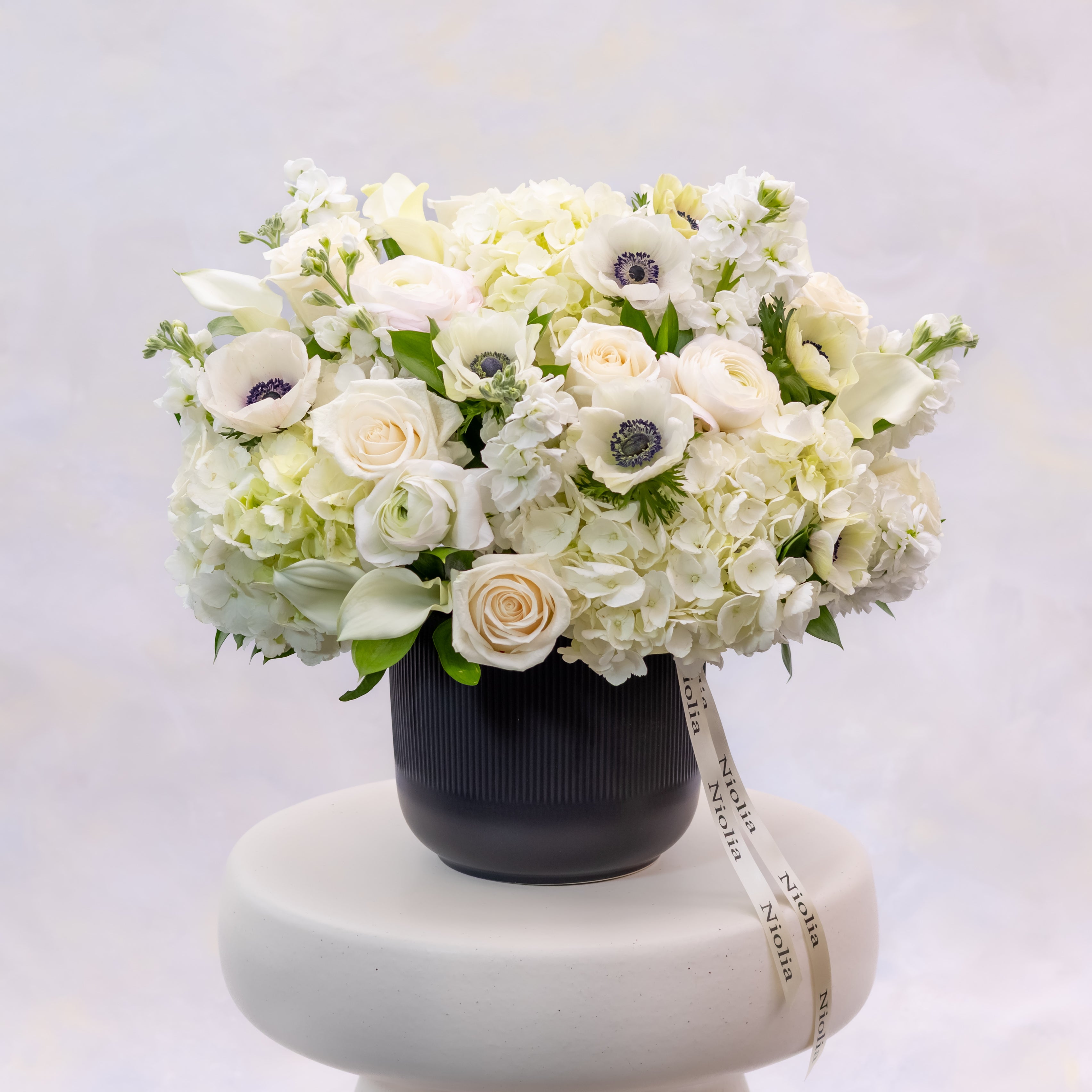 The white anemone flower arrangement in deluxe size is a balanced flower arrangement showcasing white anemones with bold dark centers in a refined black ceramic vase. Flower selection: anemones, roses, spray roses, calla lilies, hydrangeas, ranunculus, stock, and seasonal greenery. Made by Niolia Flowers in San Francisco.