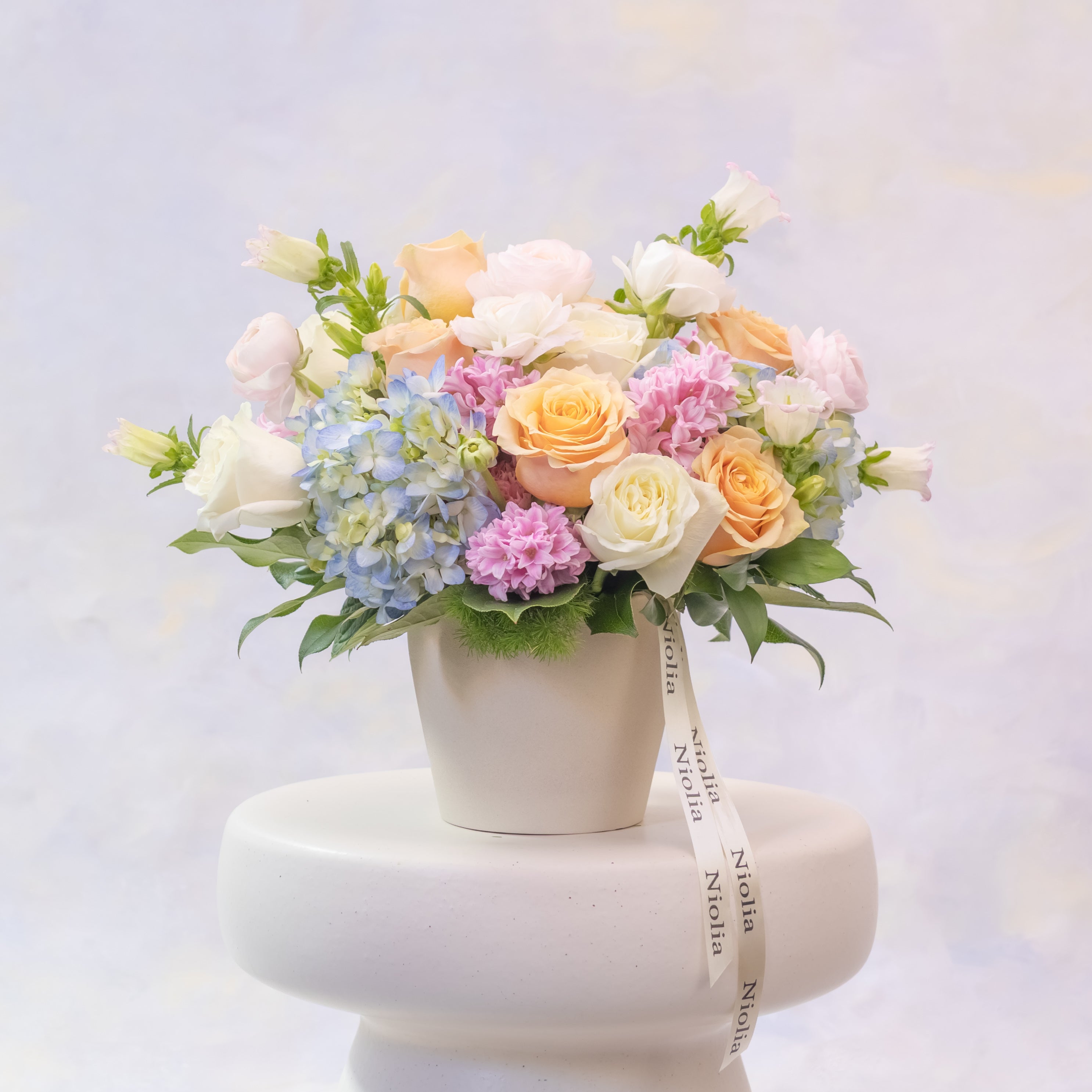Pastel-colored flower arrangement in a white pot on a white pedestal with a blurred background