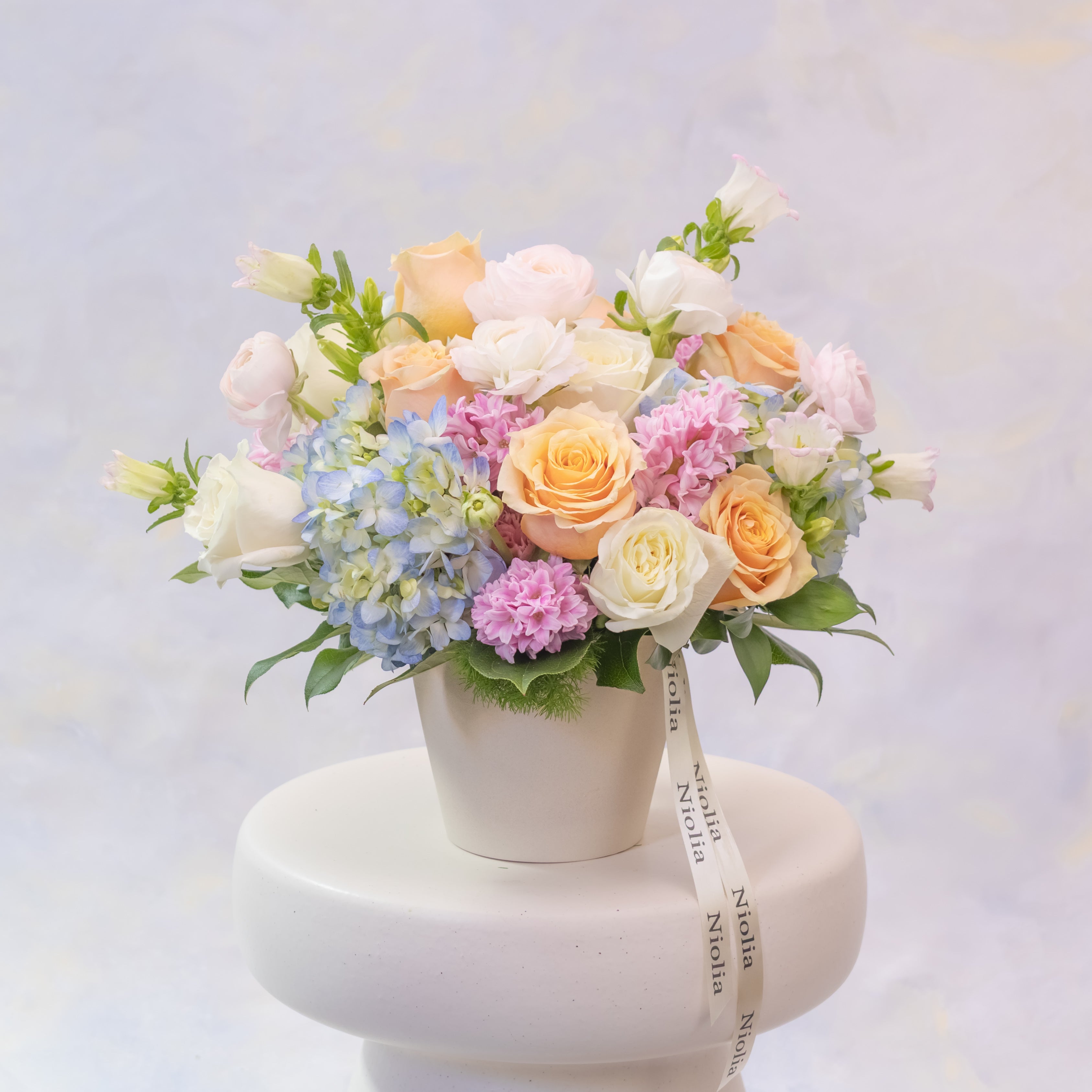 Pastel-colored flower arrangement in a white pot on a white pedestal with a light purple background.