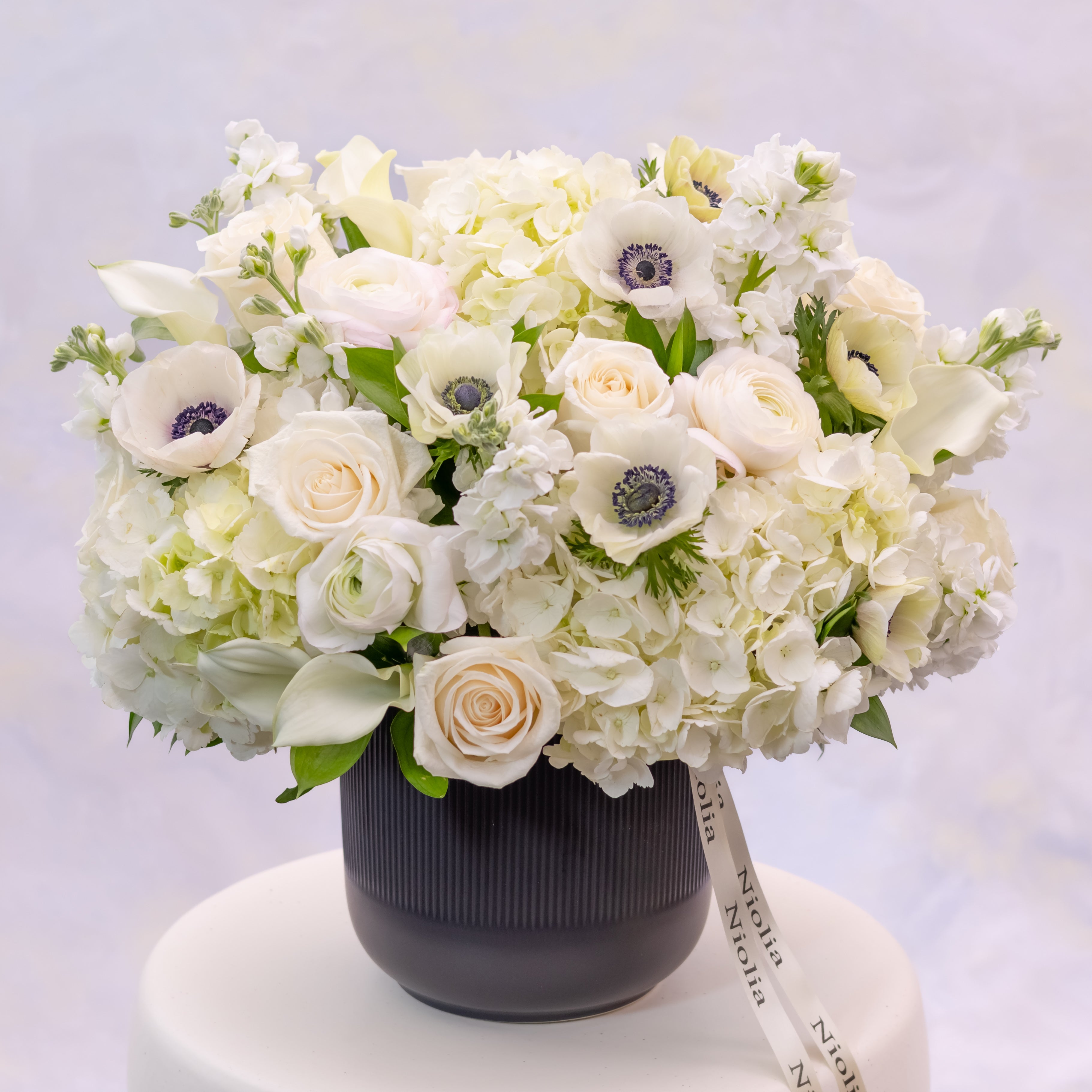 The white anemone flower arrangement in deluxe size is a balanced flower arrangement showcasing white anemones with bold dark centers in a refined black ceramic vase. Flower selection: anemones, roses, spray roses, calla lilies, hydrangeas, ranunculus, stock, and seasonal greenery. Made by Niolia Flowers in San Francisco. 