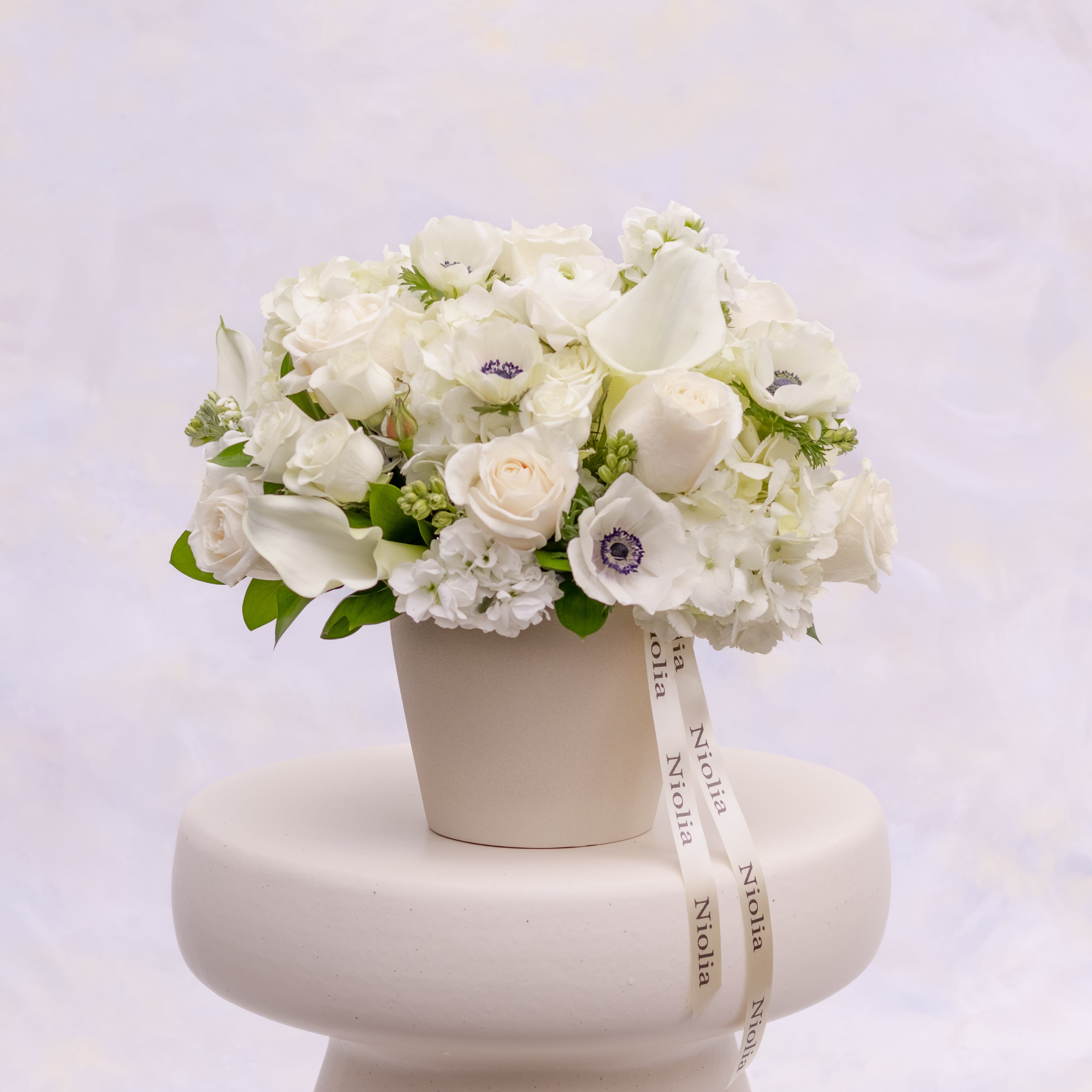 A refined white garden flower arrangement designed in a soft, natural palette of white and ivory flowers. Flower selection: anemones, calla lilies, roses, spray roses, stock, hydrangeas, and seasonal greenery. Arranged in cream ceramic vase. Made by Niolia Flowers in San Francisco.